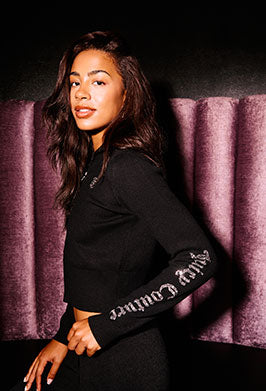 Woman wearing a black tracksuit with text on sleeves reading "Juicy Couture" against a purple velvet background