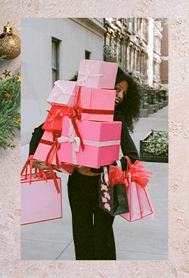 GIF of woman in black tracksuit holding a stack of pink and red gift boxes and bags standing outside, on top of a textured pink background with a pine tree and gold ornaments at the corner,