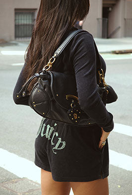 A woman from behind wearing a black long-sleeve top and black shorts with 'Juicy' written in green script, carrying a black studded shoulder bag with gold hardware.