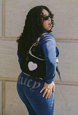 A woman with long curly black hair and sunglasses, wearing a blue long-sleeve top and matching blue pants with silver rhinestone lettering on the back, carrying a black shoulder bag with a white heart, posing against a beige stone wall.