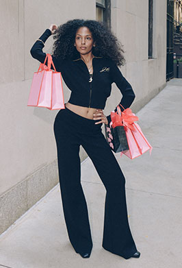 Woman in black tracksuit with a cropped zip up and flare pants holding pink gift bags while standing outside.