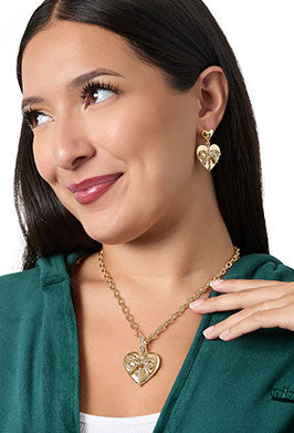 A woman wearing a gold heart pendant necklace and matching heart drop earrings, styled with a dark green top against a white background.