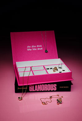 A pink jewelry display box and book set on a pink reflective surface against a black background. The open box contains several gold-tone necklaces and earrings, with additional jewelry placed on the book and outside the box.