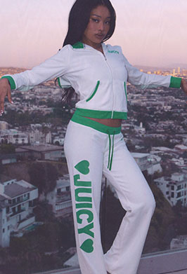 A woman modeling a Juicy Couture white and green tracksuit with 'JUICY' printed vertically on the pants, posed against a city skyline at sunset.
