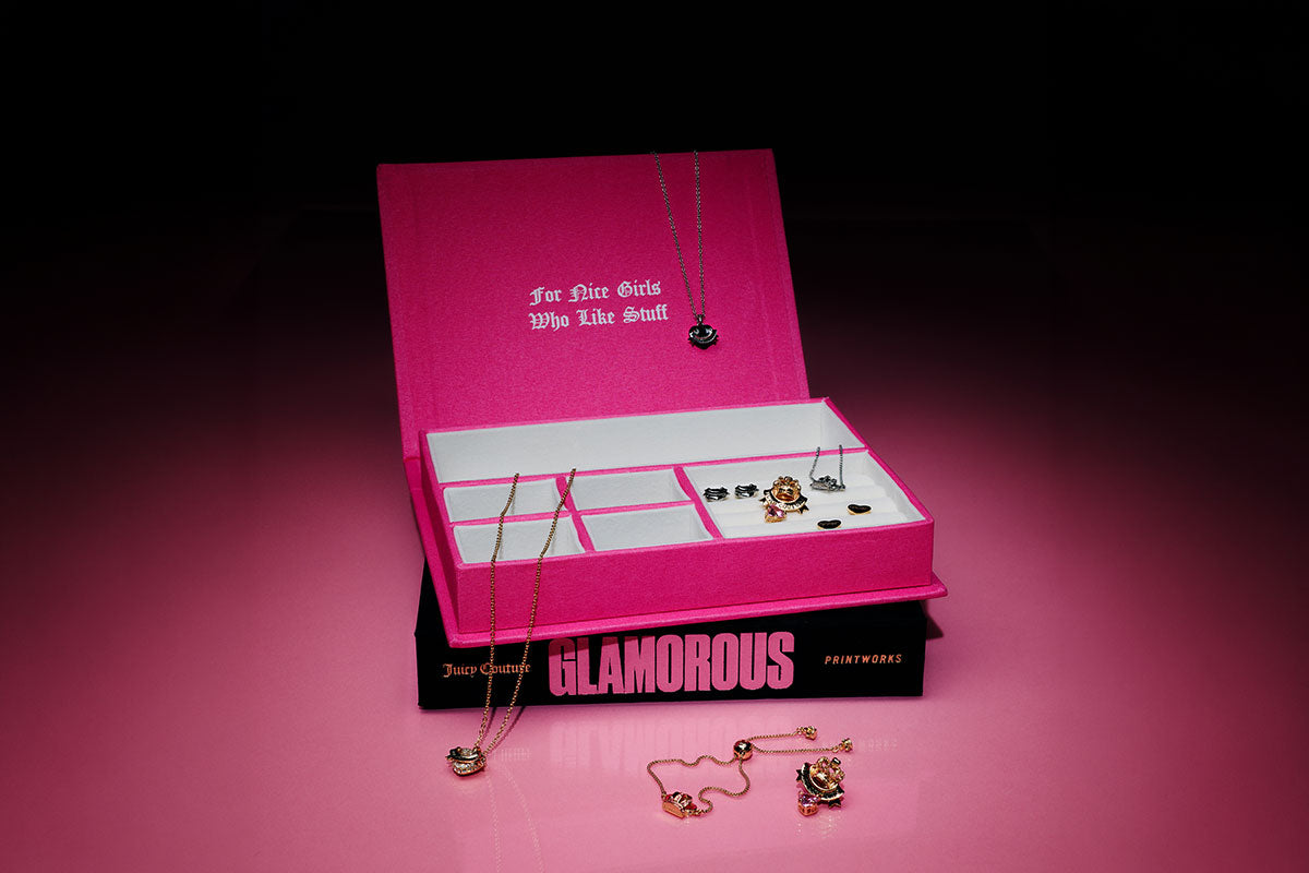 A pink Juicy Couture branded jewelry box displayed open on top of a black 'GLAMOROUS' book, with several gold necklaces and earrings arranged inside and around it against a dark pink studio background.