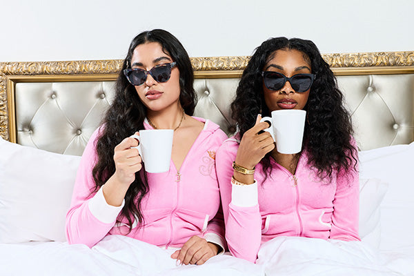 Two women in sunglasses and matching pink tracksuits holding cups in a bedroom setting