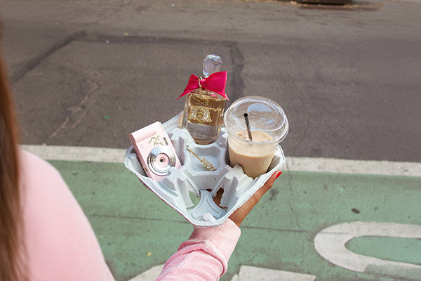 Person holding a a perfume bottle, a drink, and a pink camera in a drink tray on a street.