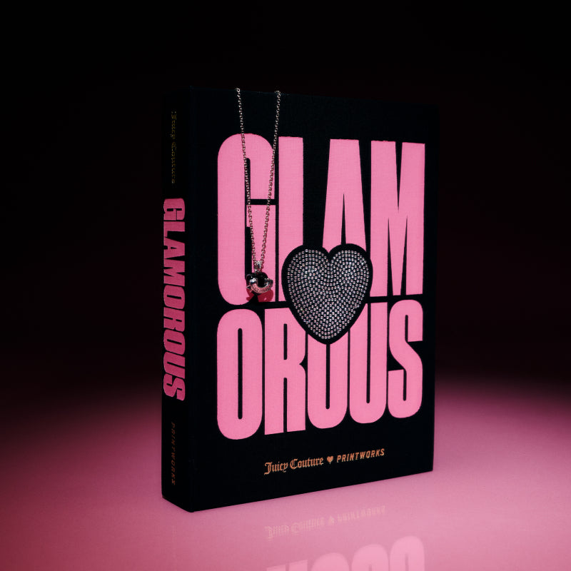 A book titled GLAMOROUS in large pink letters, featuring a heart on the cover, stands upright on a pink surface—capturing the chic allure of Juicy Couture. Draped gracefully over the book is a silver necklace with a pendant, evoking the precious finds inside the Juicy Couture x Printworks Jewelry Box. Dramatic lighting and a dark background lend an extra layer of sophistication to this jewelry box-inspired scene.