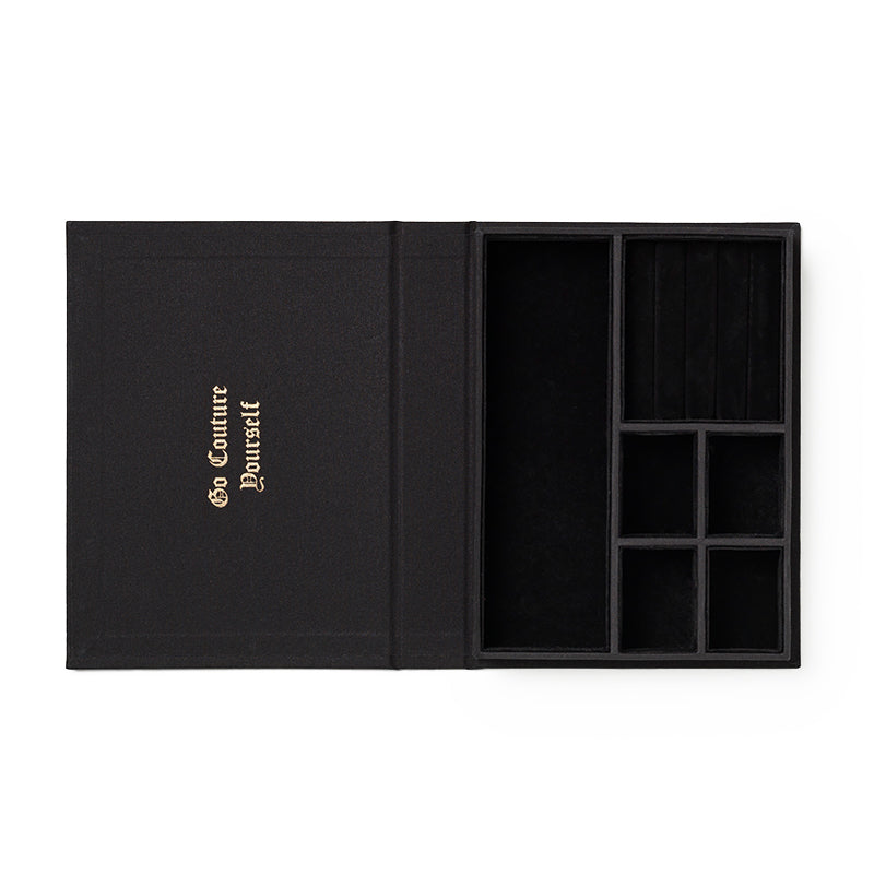 The Juicy Couture x Printworks Jewelry Box is shaped like a book and features a sleek black design. When opened, it reveals several velvet-lined compartments for glamorous jewelry storage, with “Go Couture Yourself” embossed in gold lettering on the left side cover.