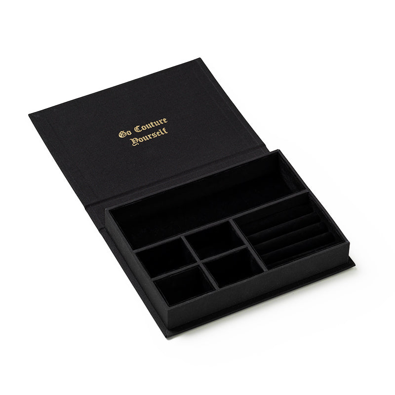 The Juicy Couture x Printworks Jewelry Box is a black jewelry box designed with multiple compartments and an open-book style, perfect for storing elegant jewelry. The lid showcases gold lettering that reads “Go Couture Yourself,” reflecting Juicy Couture’s signature flair. This chic Glamorous Jewelry Box is shown empty on a white background.