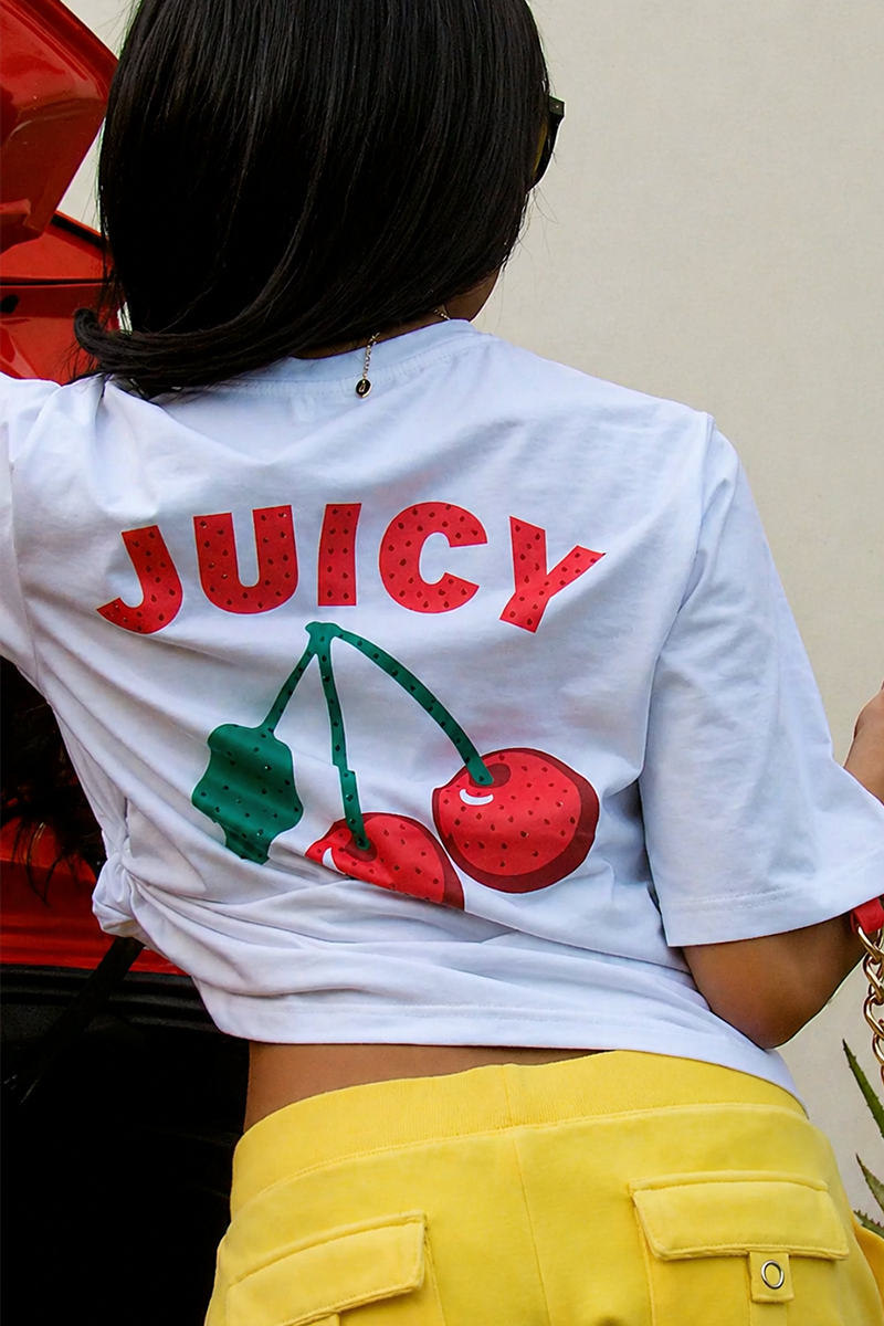 Wearing the Cherry Bling Graphic Tee featuring "JUICY" and cherries on the back, a person with long dark hair stands by a red car. They style the white tee with yellow pants for a vibrant, relaxed look.