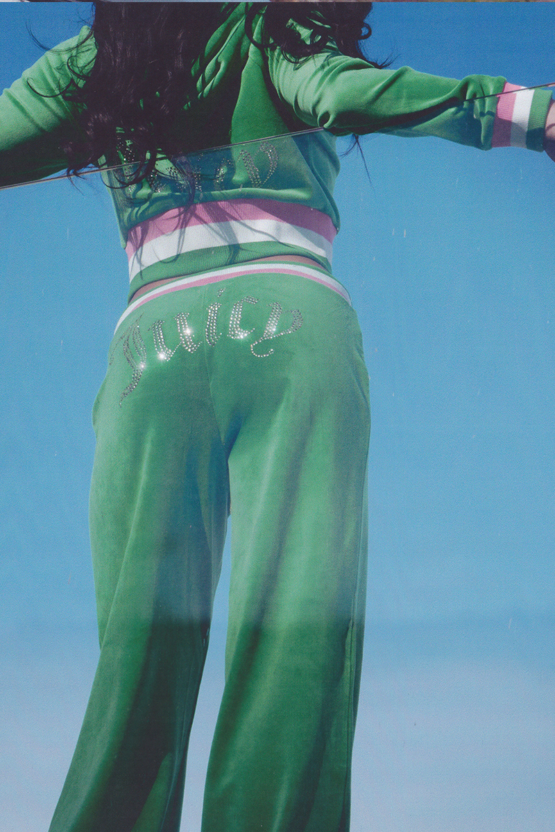Wearing Heritage Stripe Bling Velour Track Pants, which feature a striped waistband and rhinestone Juicy logo across the back, a person stands against a clear blue sky with arms outstretched, showing only their lower back and legs.