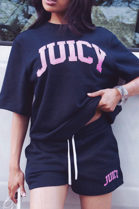A person outdoors lifts the hem of a black Collegiate Sweatshirt featuring "JUICY" in pink collegiate-style letters, paired with matching shorts.
