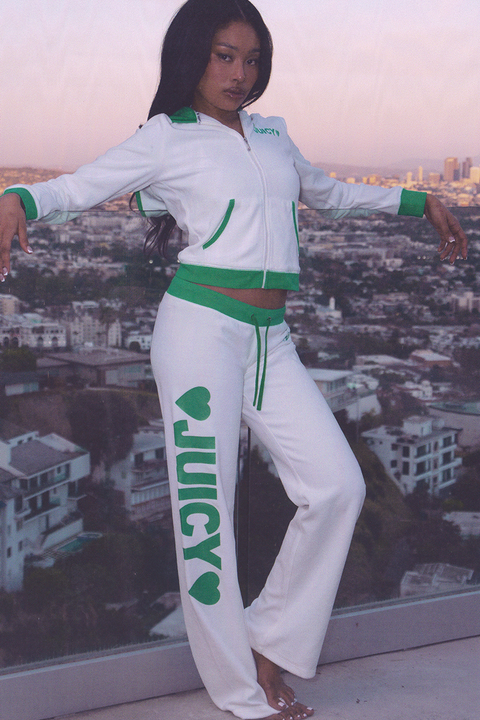 A woman poses on a balcony in the Contrast Low Rise Track Pants—white with green trim and “JUICY” plus hearts in green. The sporty loungewear contrasts with the cityscape and greenery visible in the background.