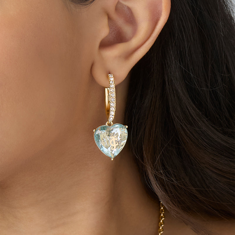 A close-up of an ear wearing the Heart Charm Hoop Earrings, featuring gold hoops embellished with small diamonds and a large, heart-shaped clear gemstone pendant. Part of the person’s dark hair and skin forms the background, allowing these striking statement earrings to stand out beautifully.