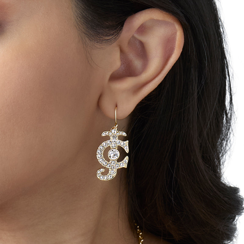 Close-up of a person’s ear adorned with the Rhinestone Logo Drop Earrings, featuring a gold Chanel-inspired logo encrusted with sparkling rhinestones and a prominent central gem. The person’s dark hair and light brown skin perfectly showcase this striking statement accessory.