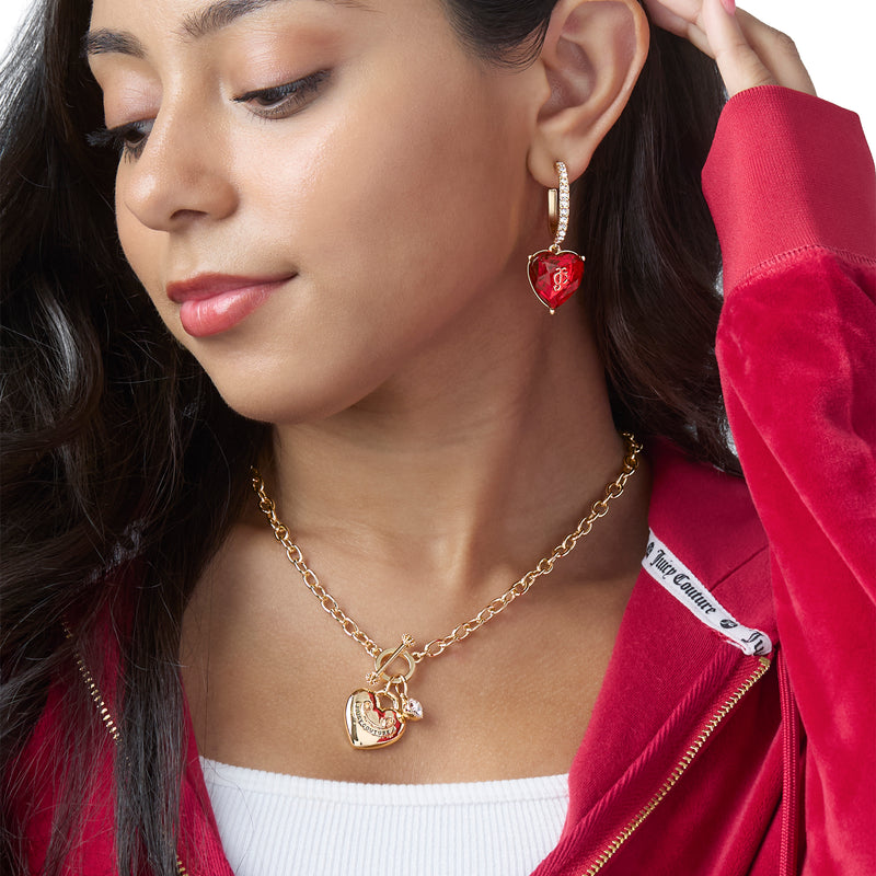 A woman in a red velvet hoodie wears Juicy Couture Heart Charm Hoop Earrings, featuring gold heart charms with a red gem, and a matching gold heart locket necklace. She looks down with a slight smile, her hand gently touching her earring.