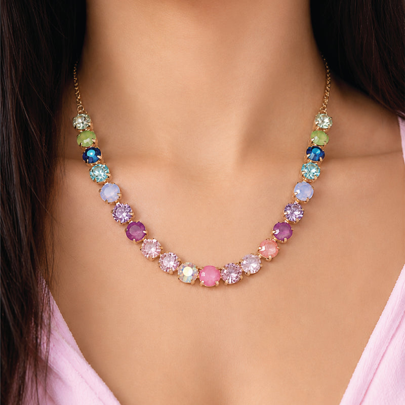 A woman wears the Tennis Necklace with Multicolor Crystals by Juicy Couture, showcasing vibrant blue, pink, purple, green, and yellow gemstones. This eye-catching gold-plated statement jewelry piece brings a playful flair to her style as she pairs it with a light pink top against a neutral background.