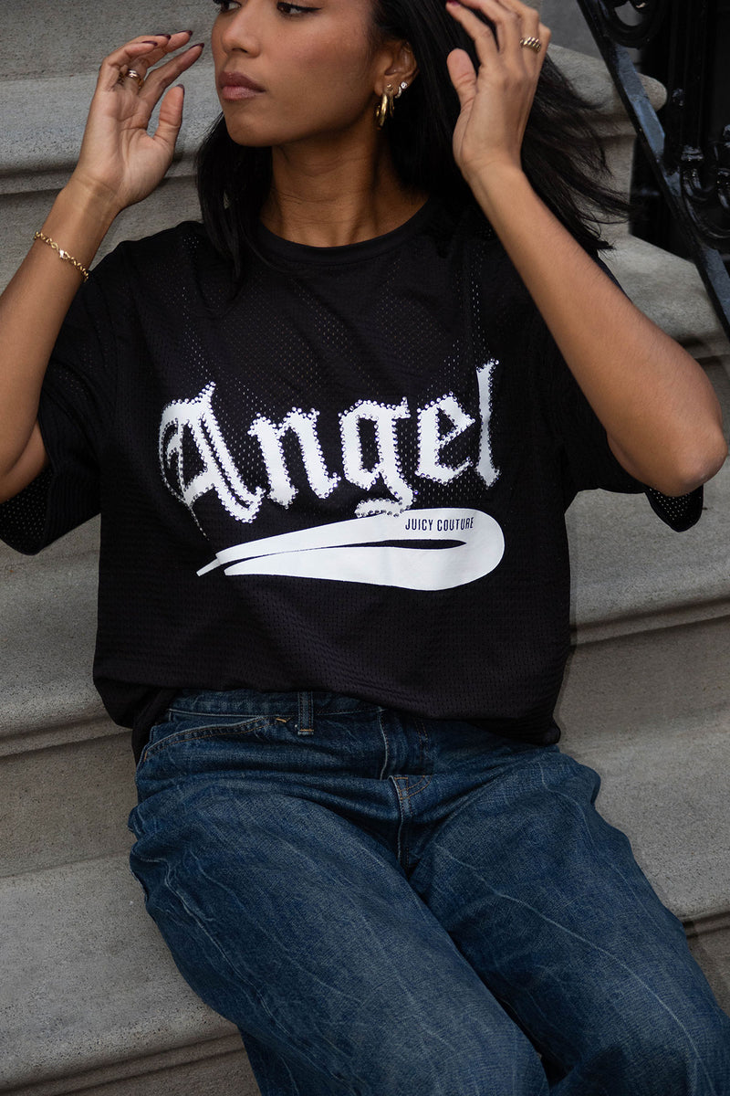 A woman wearing the Angel Couture Oversized Tee Dress in black with "Angel" printed in white, paired with blue jeans, sits on outdoor steps and raises her hands near her face.