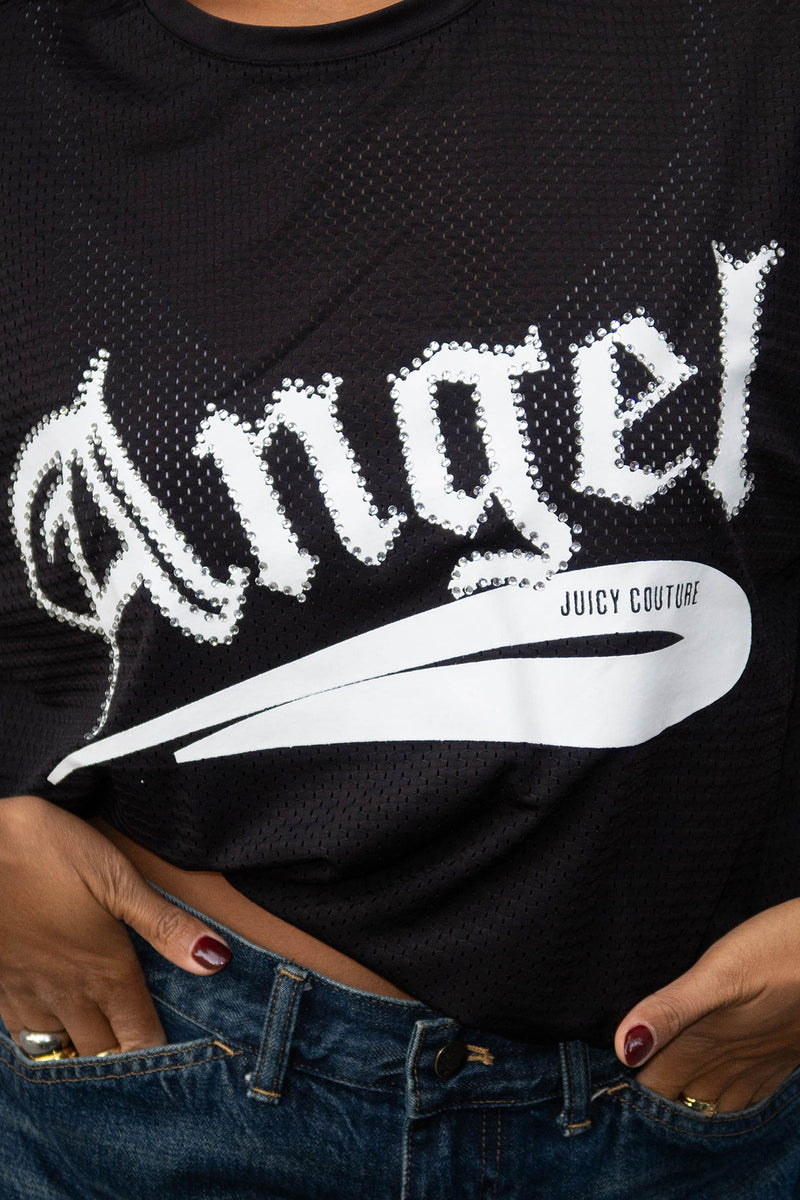 A person wears the Angel Couture Oversized Tee Dress, a black mesh piece featuring bold white “Angel” gothic letters and a white Juicy Couture swoosh, tucked into blue jeans with hands on hips for a chic, sporty style.