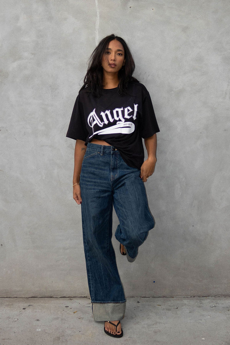 A woman with long dark hair poses confidently against a gray wall, wearing the Angel Couture Oversized Tee Dress in black with "Angel" lettering, paired with cuffed wide-leg jeans and black sandals.