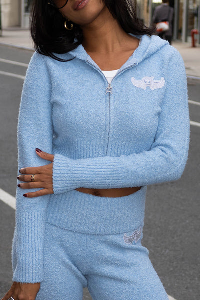 Woman in a light blue, cropped Angel Couture Knitwear Patch Hoodie and matching pants stands on a city street, arms crossed. With dark hair, sunglasses, and statement rings, she channels effortless sporty luxe style.