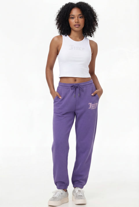 A woman with curly hair wears the Sora Graphic Jogger—REJUICED, styled with a white sleeveless crop top featuring Juicy text and white sneakers, standing with hands in pockets against a plain background.