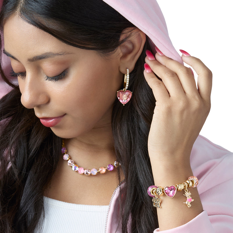 A woman wearing a pink hoodie, heart-shaped pink and gold earrings, the Juicy Girl and Scottie Dog Charm Bracelet with charms, and a matching pink necklace, looking down with closed eyes.