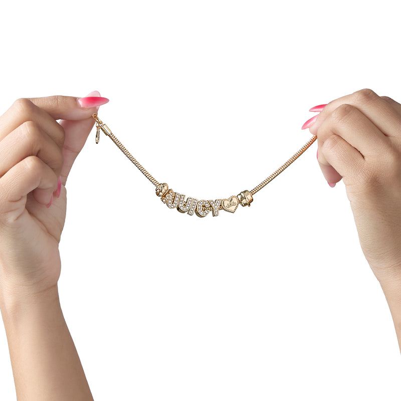 Two hands with pink nails hold the Juicy Couture Logo Charm Bracelet, featuring letter charms that spell “JUICY” adorned with sparkling rhinestones, highlighting bold statement jewelry against a white background.