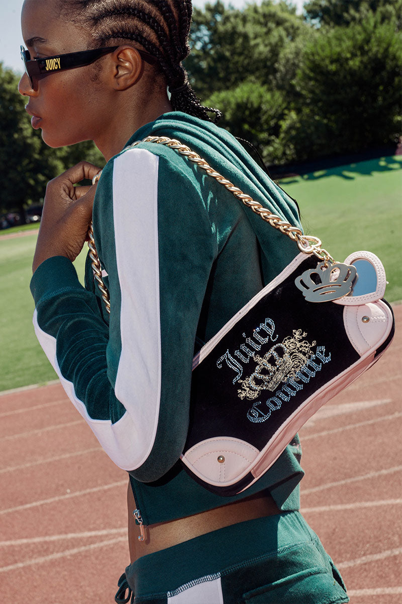 A woman outdoors on a track field wearing a green and white tracksuit and sunglasses, carrying a black and white Juicy Couture shoulder bag with a gold chain strap.