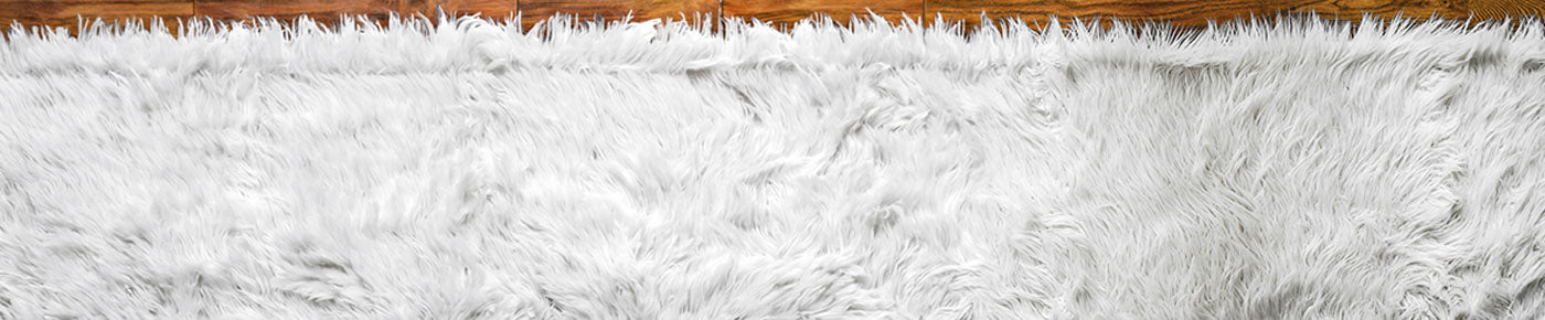 A close-up of a white shaggy faux fur rug or blanket texture with a warm brown wooden surface visible along the top edge.