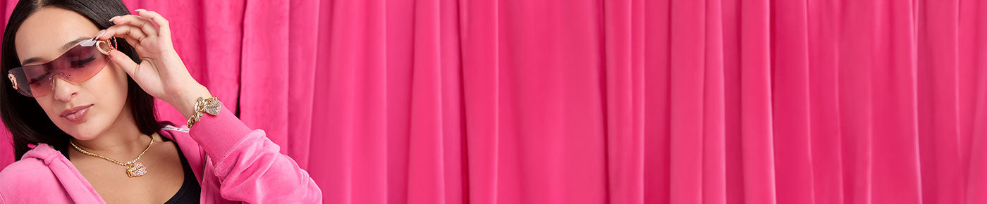 A woman wearing dark sunglasses and a pink hoodie poses against a bright pink curtain backdrop.