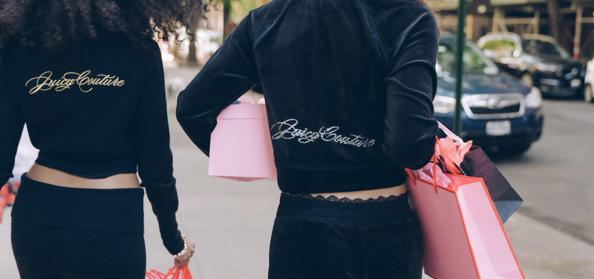 Two women from behind wearing black velvet cropped jackets with 'Juicy Couture' embroidered in gold on the back, walking on a city street and holding pink and red gift bags.