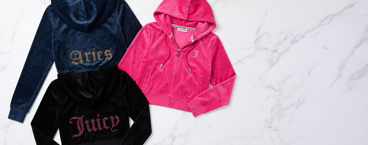 Three Juicy Couture zip-up hoodies in navy blue, black, and bright pink, laid flat on a white marble background. Each hoodie features the brand's signature embroidered gothic lettering on the back or front.