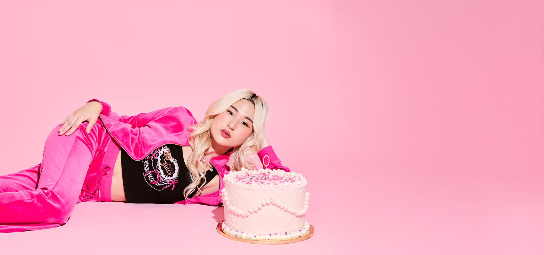 Person in pink tracksuit and black top with a cake on a pink background