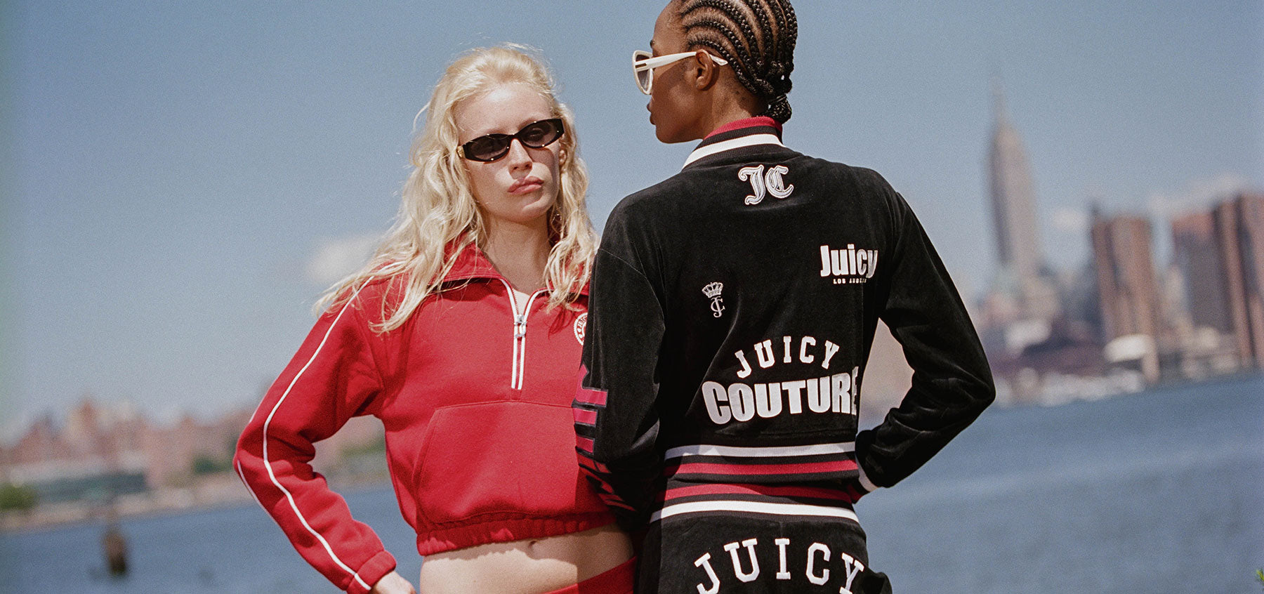 Two models wearing Juicy Couture apparel, one in a red zip-up jacket and one in a black track jacket with 'Juicy Couture' text on the back, set against a city skyline and water background.