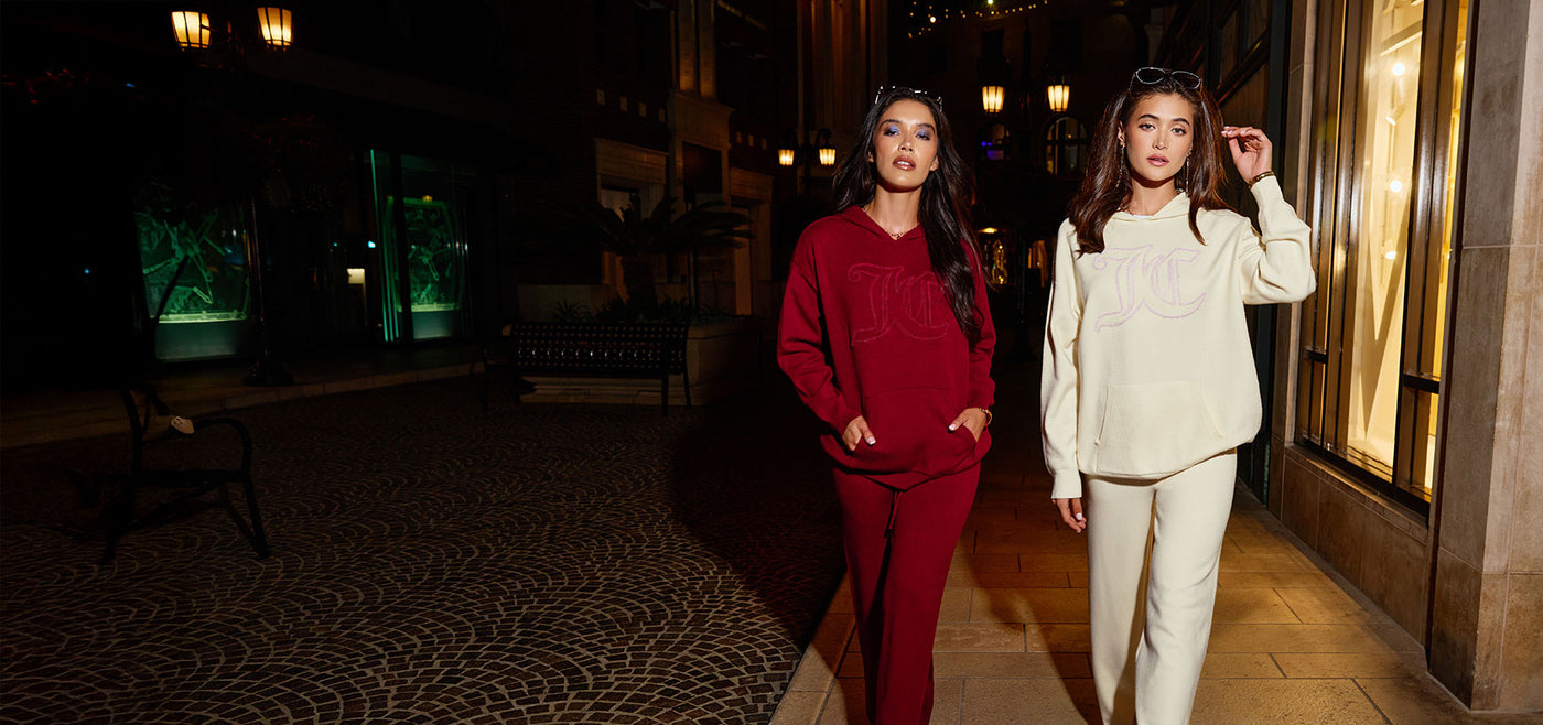 Two women in red and beige Juicy Couture tracksuits standing in a dimly lit street with decorative elements.