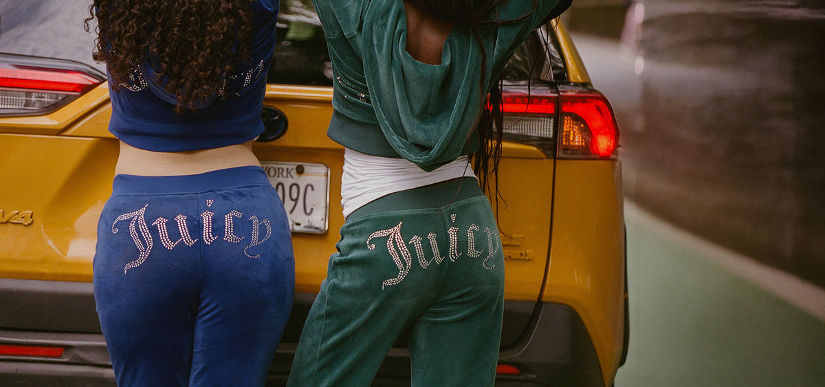 Two women standing behind a yellow car, wearing matching athletic sweatpants with 'Juicy' written across the back in a gothic font. One pair is blue and the other is green, with metallic rhinestone lettering.