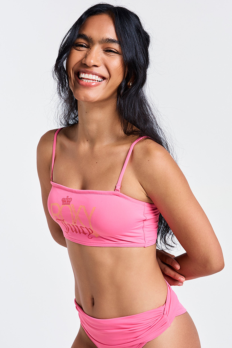 A woman with long dark hair smiles, wearing the Roxy x Juicy Bandeau Bikini Top in Azalea Pink with gold lettering, made from recycled nylon. She stands against a plain light background with her hands behind her back.