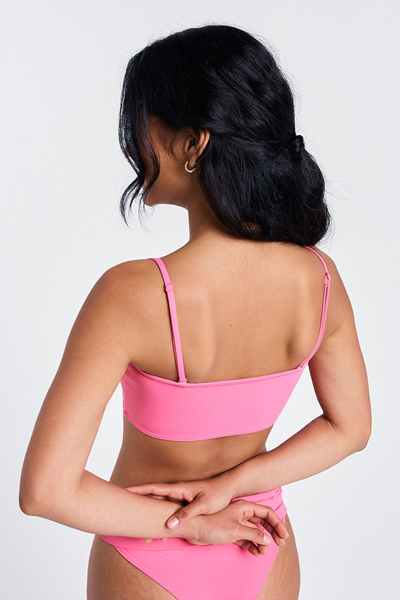 A woman with long black hair faces away, wearing the Roxy x Juicy Bandeau Bikini Top in Azalea Pink (recycled nylon), one arm bent behind her back as she poses against a plain white background.