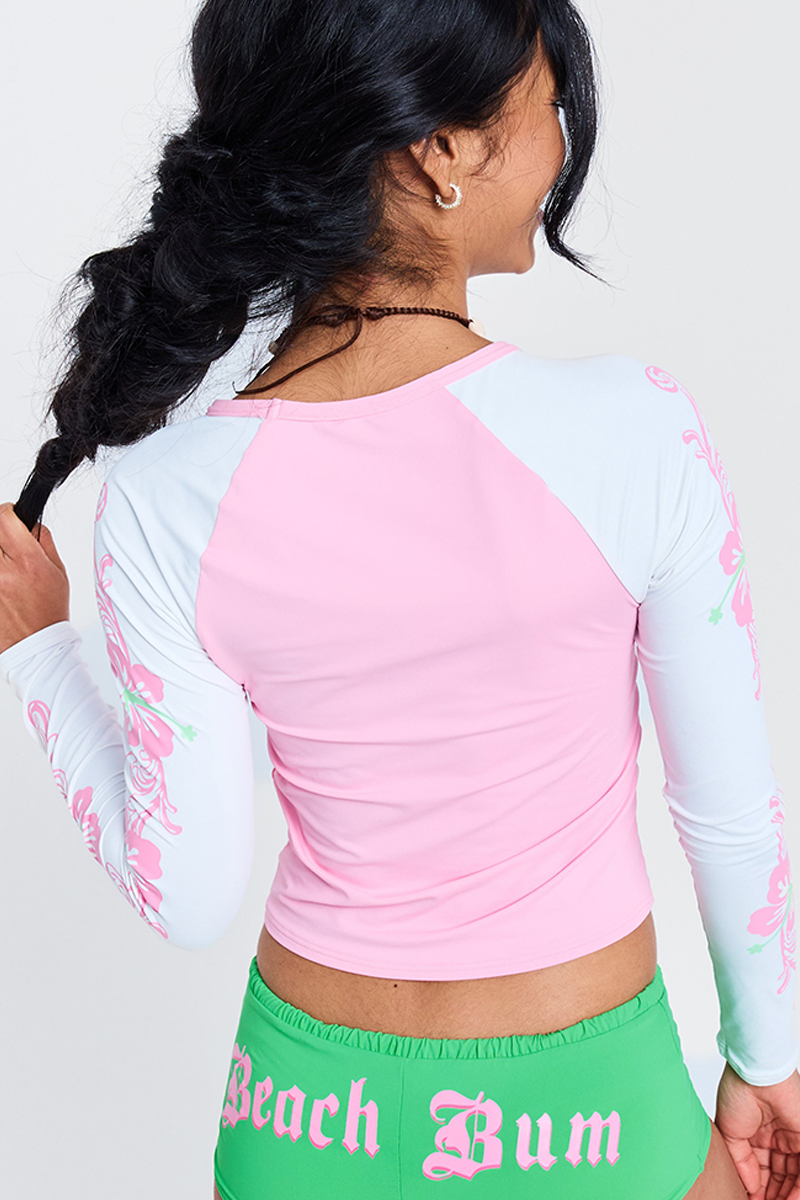 A woman with long dark hair in a ponytail faces away, wearing the Roxy x Juicy Long Sleeve Lycra Rashguard in pink and white and green shorts with "Beach Bum" printed in pink Gothic letters on the back.