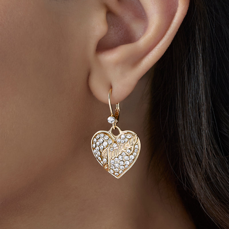 A close-up of an ear adorned with the Crystal Heart Charm Dangle Earrings—gold heart-shaped drops encrusted with sparkling clear stones and engraved in cursive, capturing the playful elegance of crystal heart jewelry.