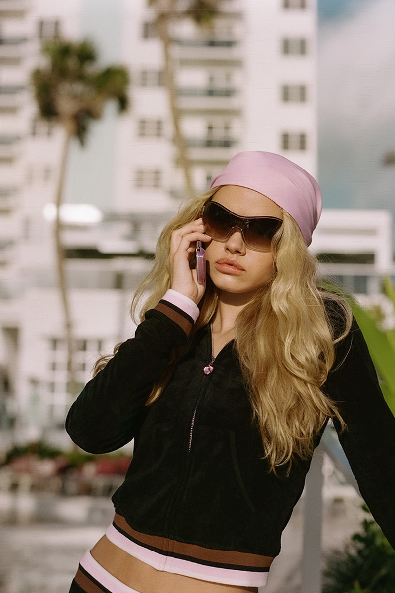 A woman with long blonde hair, wearing a pink headscarf, large sunglasses, and a black and pink Roxy x Juicy Fitted Hoodie stands outdoors on her cellphone. Palm trees and a white building are in the background.
