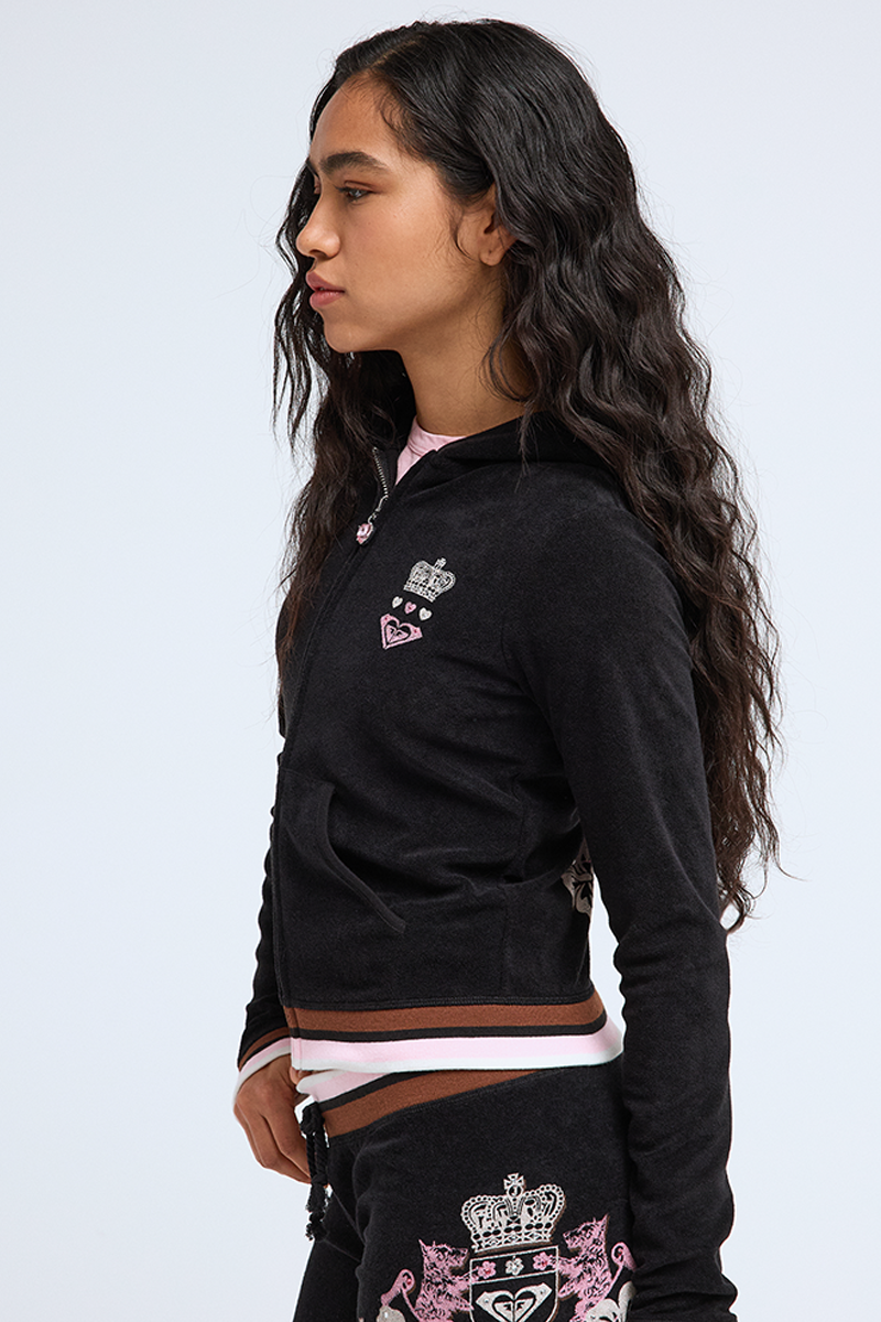A young woman with long wavy hair wears a Roxy x Juicy Fitted Hoodie with embroidered crown and heart patches, standing in profile against a plain light background.