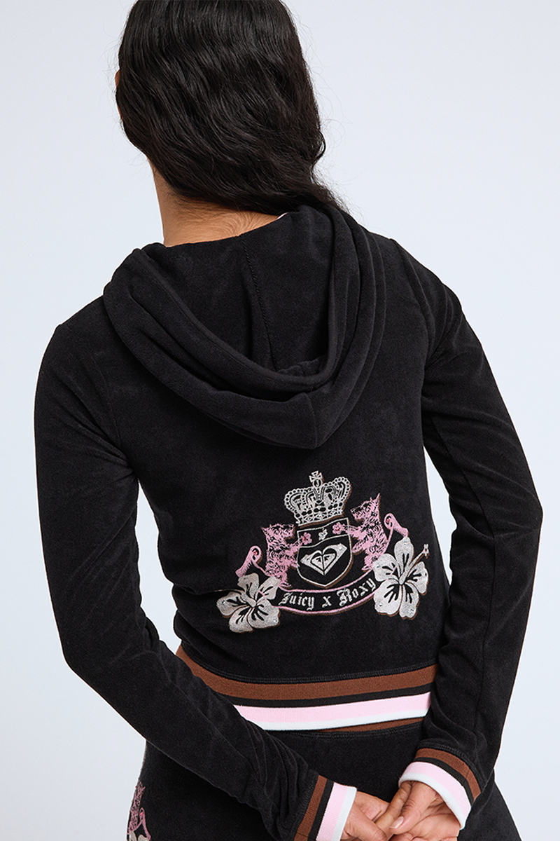 A person with long dark hair, seen from behind, wears the Roxy x Juicy Fitted Hoodie featuring pink and white floral designs, a crest, and the words “Juicy x BSR” on the back. Their hands are clasped behind them.