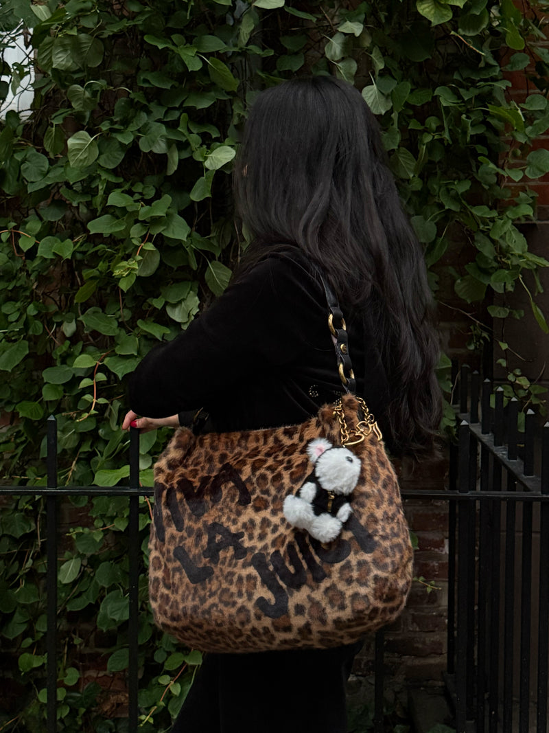 A person with long dark hair carries a leopard-print Viva La Juicy Hobo Bag with black text and a small plush panda bag charm for an everyday silhouette.