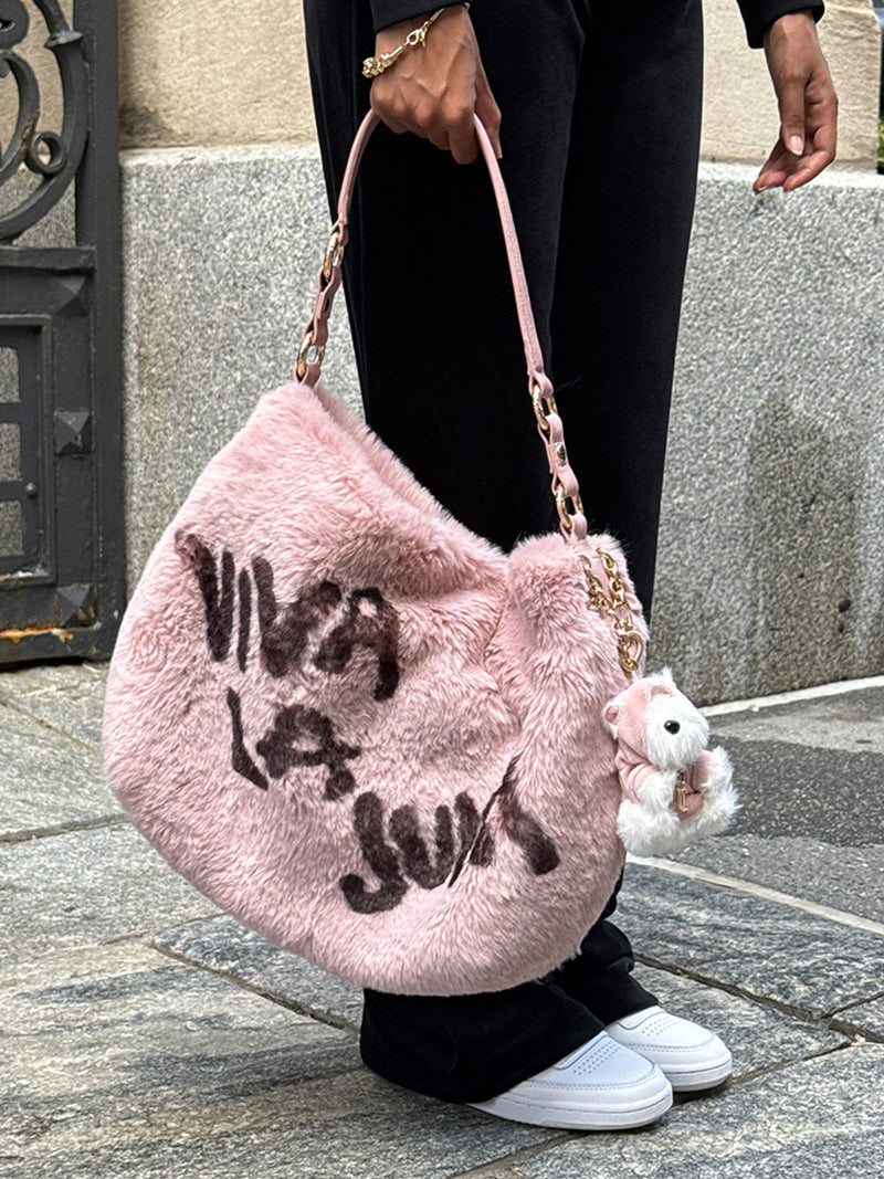 A person holding a pink faux fur Viva La Juicy Hobo Bag featuring dark script lettering and a small plush charm on the chain strap, perfect for an everyday silhouette.