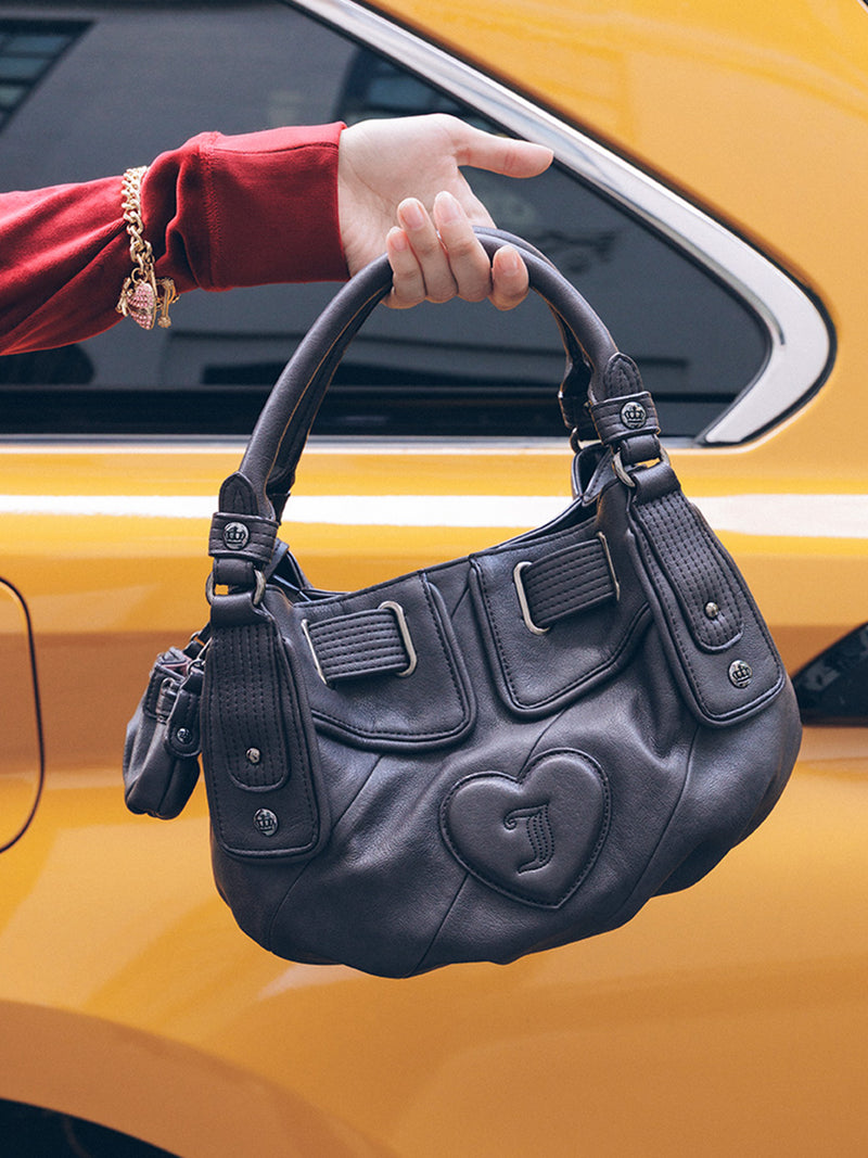 A hand holds a black Twist & Shout Crossbody Bag with a heart-shaped emblem, styled for everyday wear in front of a yellow car.