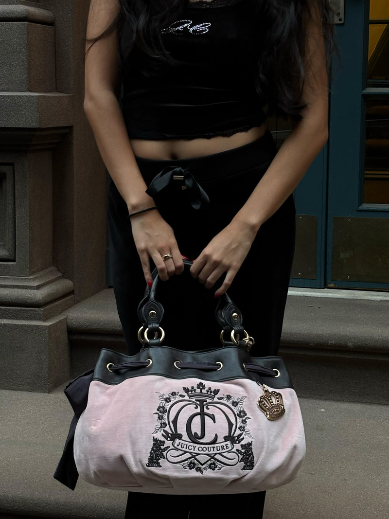 A person holds a large, light pink Crowns & Kisses Satchel Bag featuring a black embroidered logo with a crown design and a hanging metallic crown charm in a statement-making silhouette.