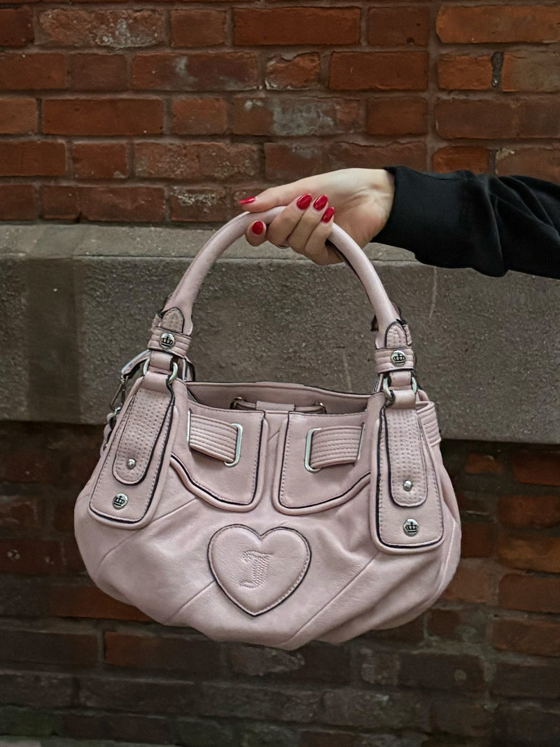 A hand holds a pale pink, slouchy Twist & Shout Crossbody Bag featuring a heart-shaped emblem on the front.